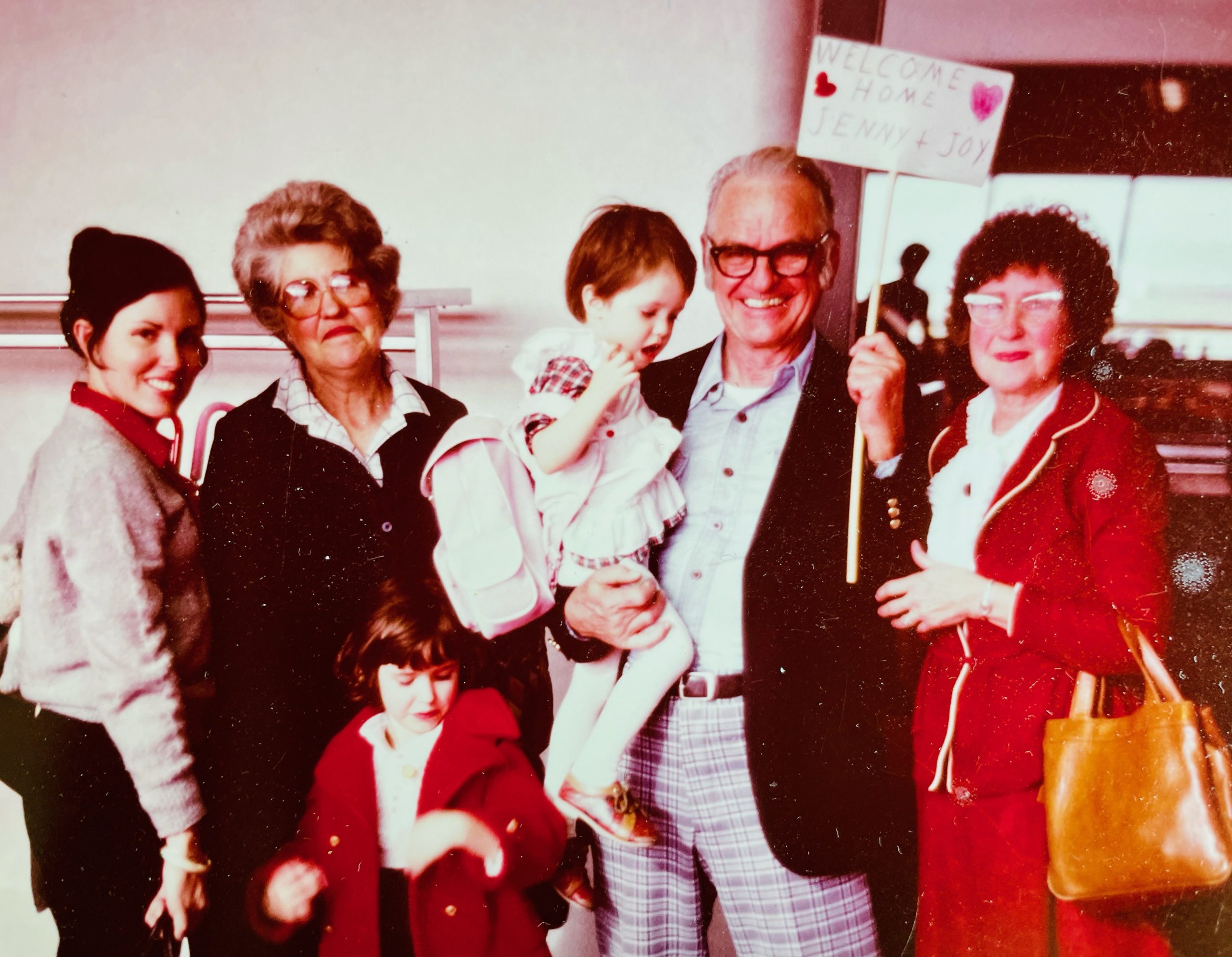 vintage photo of family meeting at airport, grandfather holds a baby, girl in red coat smiles alongside grandmother and older sister.